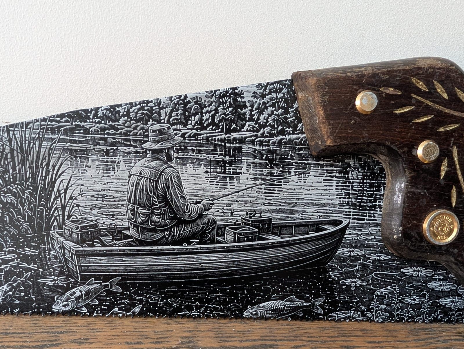 A man fishing from a rowing boat on a lake, engraved along an old hand saw